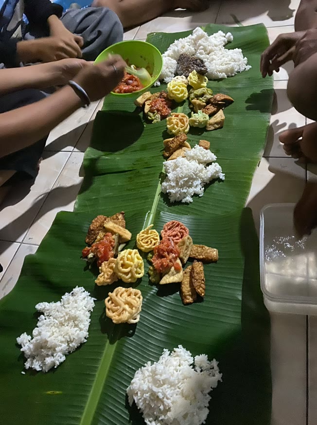 A long banana leaf laid on the floor with mounds of rice, fried tempeh, tofu and sambal, with several hands reaching in to eat together