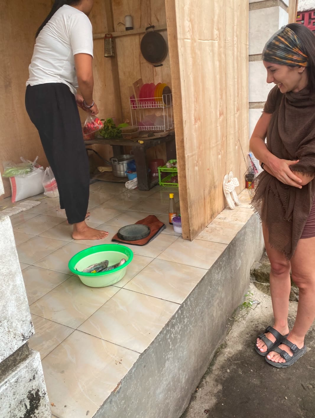 Laura laughing at a green basin of whole fresh fish on the kitchen floor while her host prepares vegetables at the bench