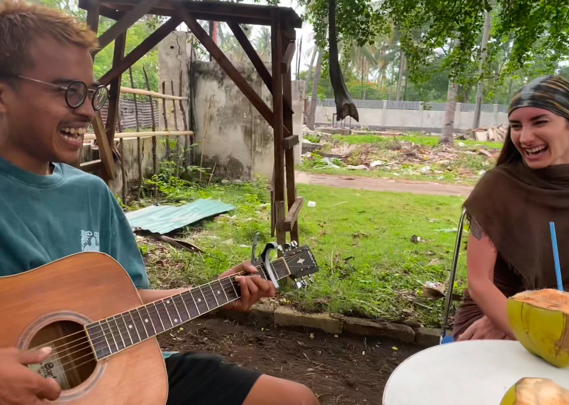 A young Indonesian man playing acoustic guitar and laughing with Laura after dinner, fresh coconuts on the table between them