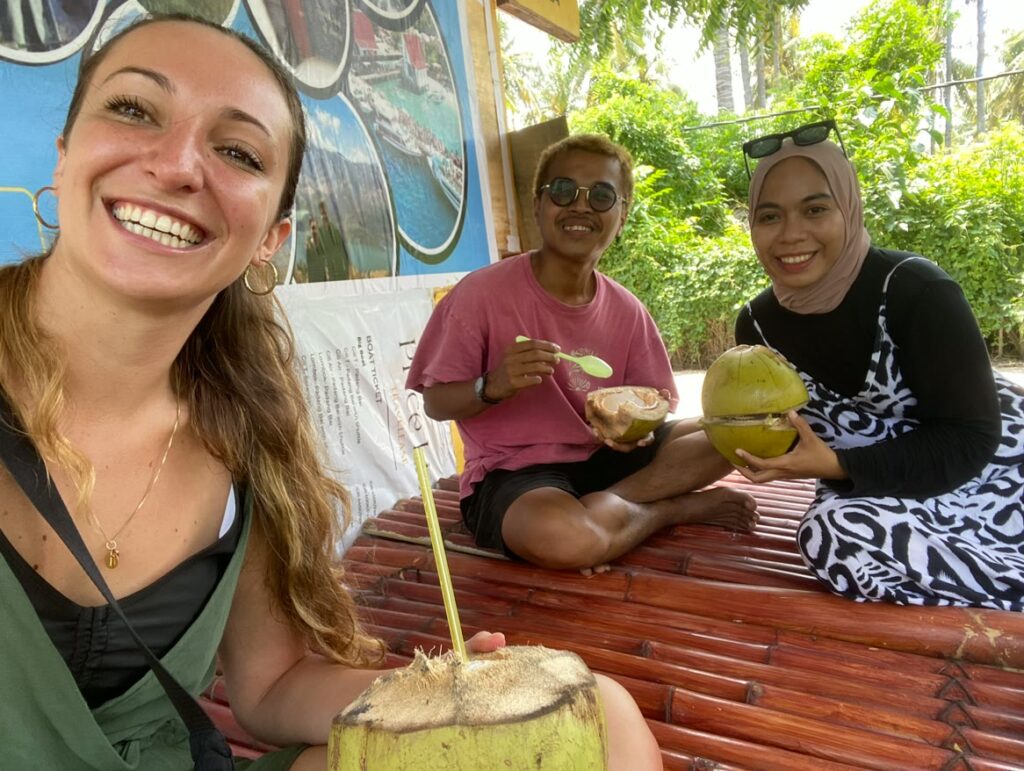 Laura sitting on a bamboo bench with her Indonesian hosts, all three drinking from fresh green coconuts and smiling