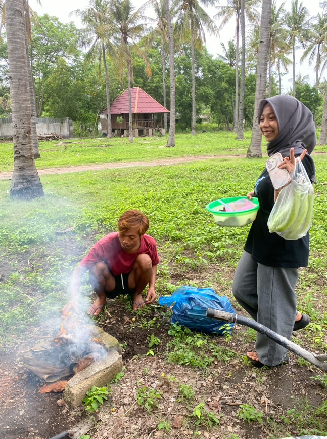 A young woman carrying vegetables and a bowl of prepared fish smiles at the camera while a man lights a coconut husk fire, with a traditional Sasak rice barn behind