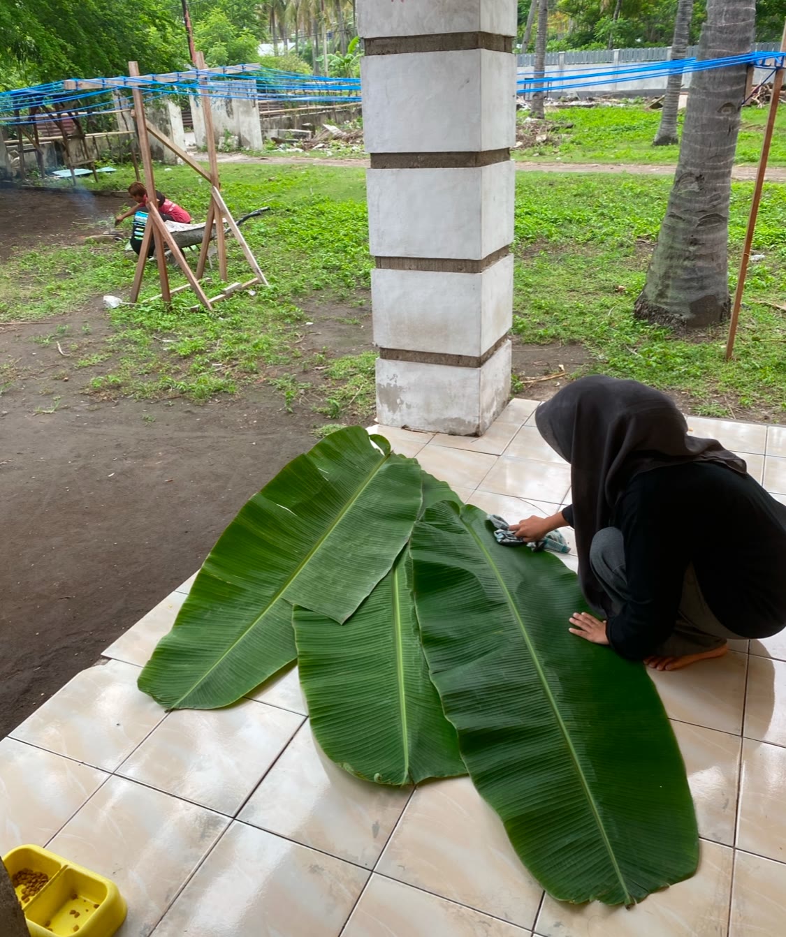 A woman kneeling on the floor wiping large banana leaves clean, which will be used in place of plates for dinner