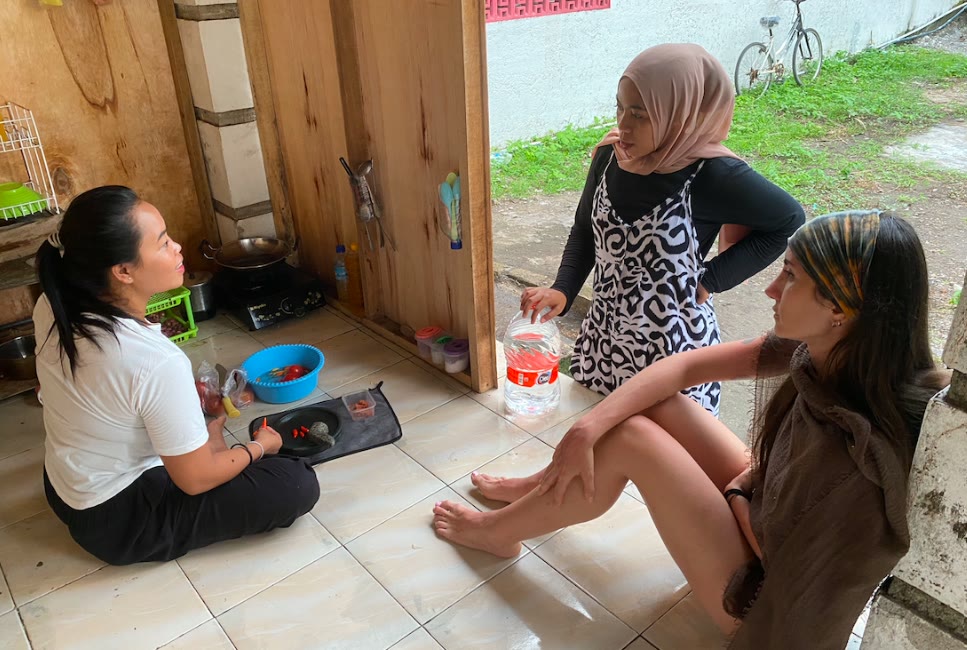 Three women sitting on the tiled floor of a family kitchen in Lombok, sorting fresh red chillies into a bowl and talking